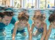 Drei Kinder und ein Schwimmlehrer tauchen ohne Schwimmbrillen gemeinsam in einem hellen Schwimmbecken unter.