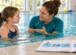 Eine Schwimmlehrerin erklärt einem Kind im Wasser eine Übung. Am Beckenrand liegt ein geöffnetes Schwimmheft mit illustrierten Lernschritten.