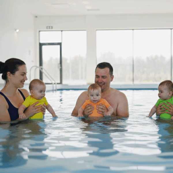 2 Frauen und 1 Mann beim Babyschwimmen im Hallenbad. Sie stehen bis zur Brust im Wasser und halten ihre Babies in sicherem Griff vor sich. Alle sind entspannt.