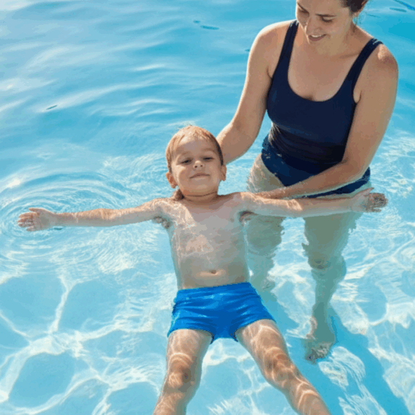 Basiskompetenzen lernen: Ein kleiner blonder Junge schwebt auf dem Rücken im Wasser, mit ausgebreiteten Armen und Beinen. Seine Mutter hält stützend die Hände unter seinem Körper.